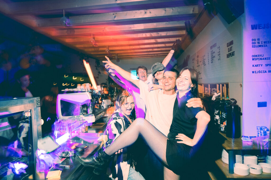 Lifestyle photography by Łukasz Zając, group of friends posing and celebrating together near a bar counter with vibrant colorful lights, Kraków, Poland