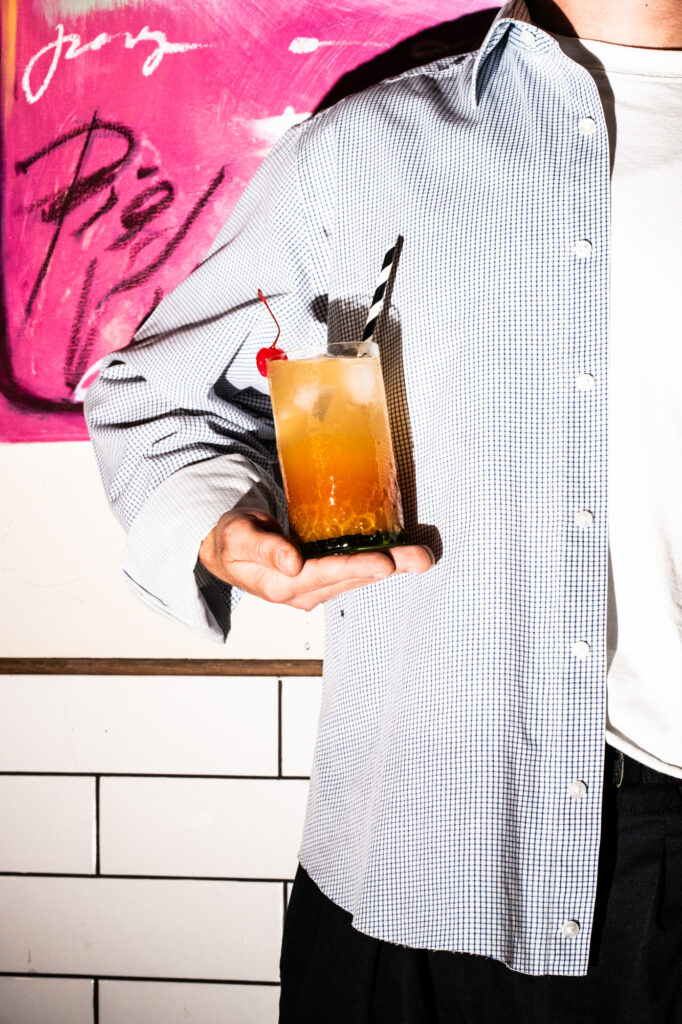 Commercial beverage and brand photography by Łukasz Zając, person holding a cocktail in a checkered shirt against pink graffiti and white tiles background, Poland