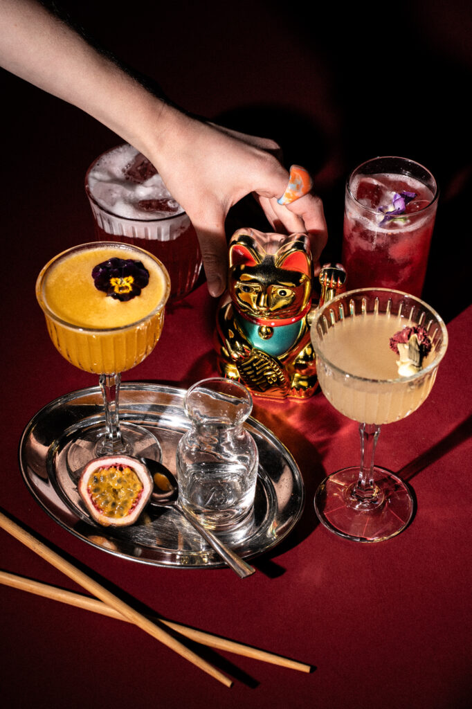 Commercial beverage and cocktail photography by Łukasz Zając, colorful drinks with golden lucky cat figurine on a dark red background, Kraków, Poland