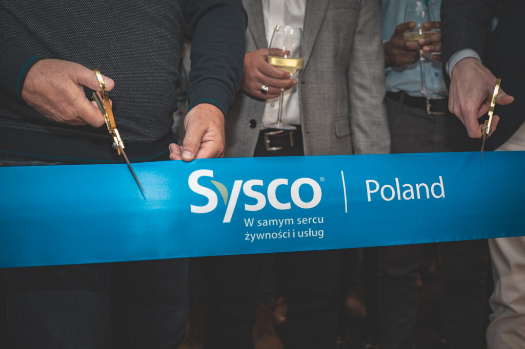Ribbon-cutting ceremony featuring officials or executives cutting a blue ribbon that reads "Sysco Poland".