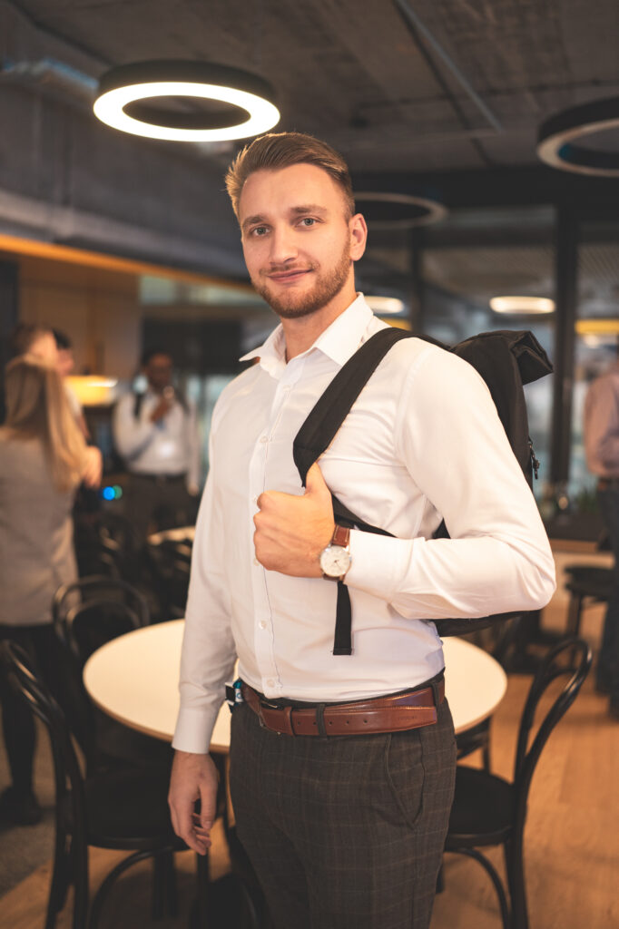 Portrait of a young man smiling while wearing a white shirt and a bag strap over his shoulder at a corporate event or office setting.