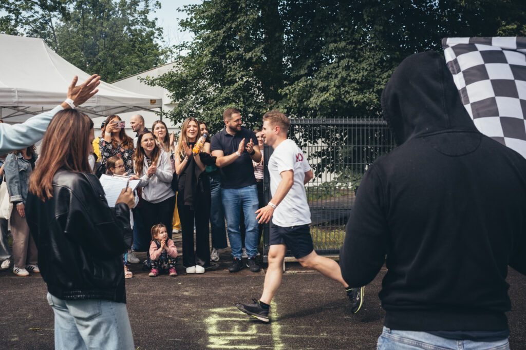 An outdoor event scene showing a crowd of people watching and cheering as someone runs towards a finish line with a checkered flag nearby.