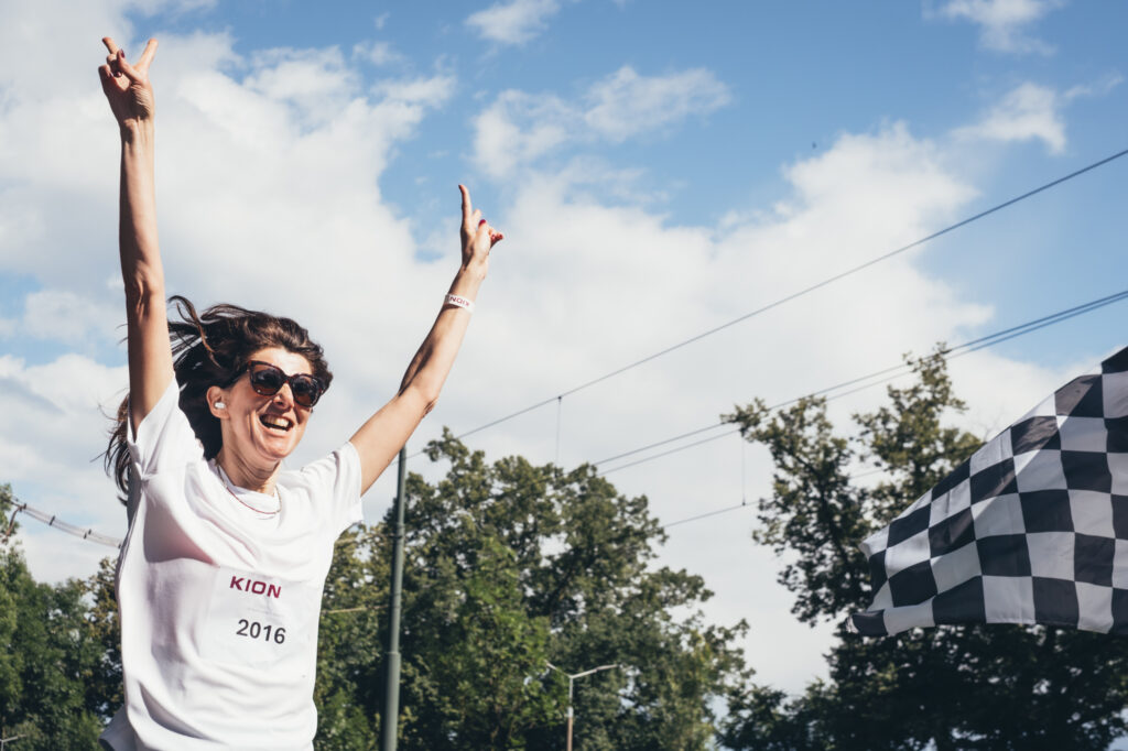 A joyful woman with her hands raised in celebration at an outdoor event or race under a cloudy sky.