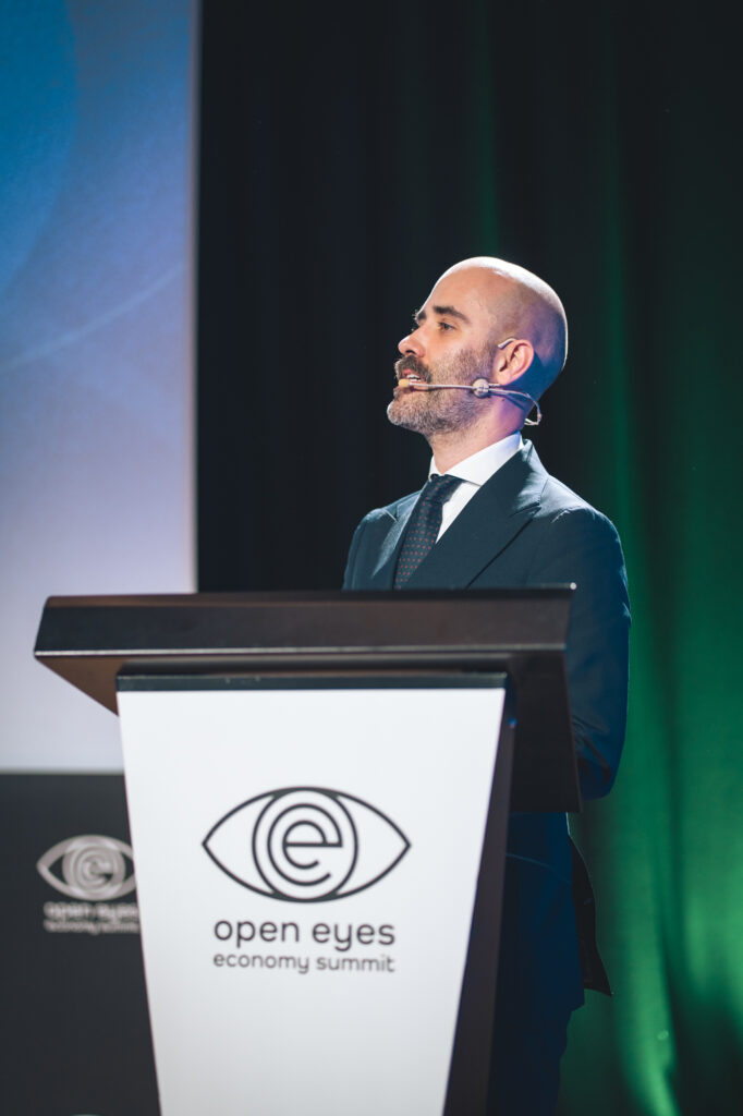 A speaker presenting at a podium bearing the Open Eyes Economy Summit branding.