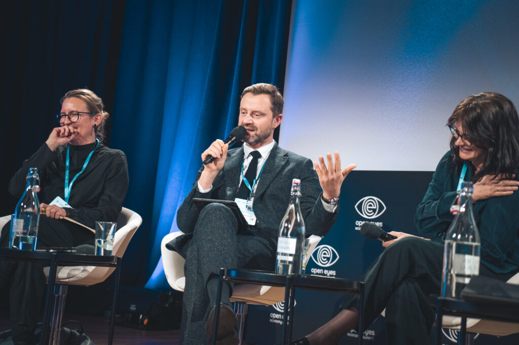 Panel discussion with speakers on stage seated in front of a blue and white backdrop during a conference.