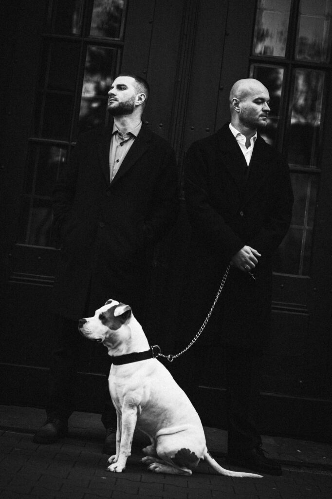 Commercial portrait photography by Łukasz Zając, two men in dark coats with a white dog on a leash in front of a building, Poland