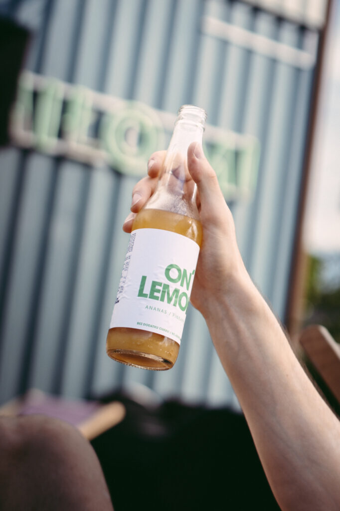 Commercial and lifestyle product photography by Łukasz Zając, hand holding up a bottle of beverage outdoors against an urban backdrop, Poland