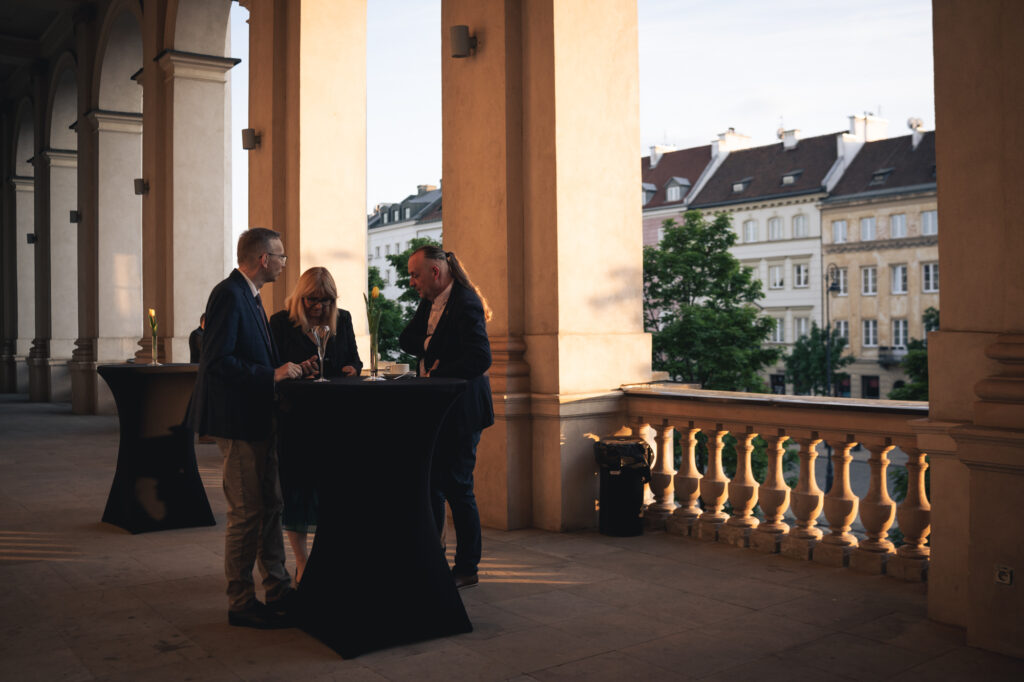 People networking outdoors on a balcony or terrace with columns, overlooking a cityscape during golden hour.