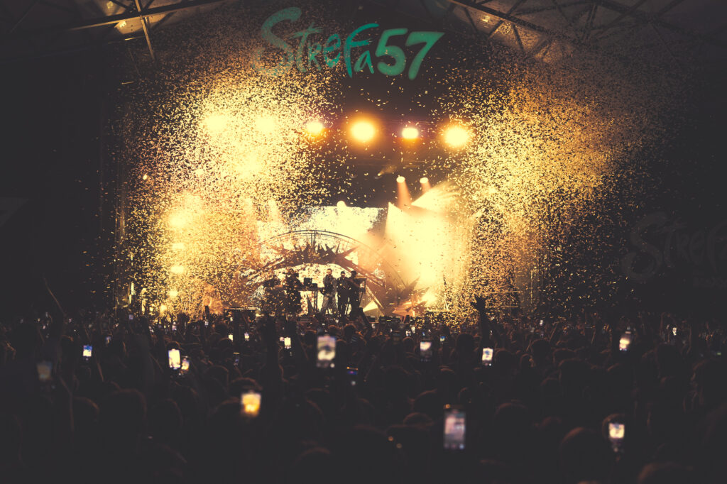 Culture and events concert photography by Łukasz Zając, wide shot of a crowded venue with stage lights and particles in the air, Poland