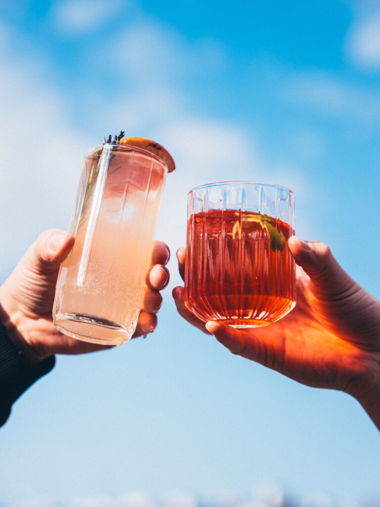 Commercial beverage and lifestyle photography by Łukasz Zając, two people toasting with cocktail glasses against a blue sky, Kraków, Poland