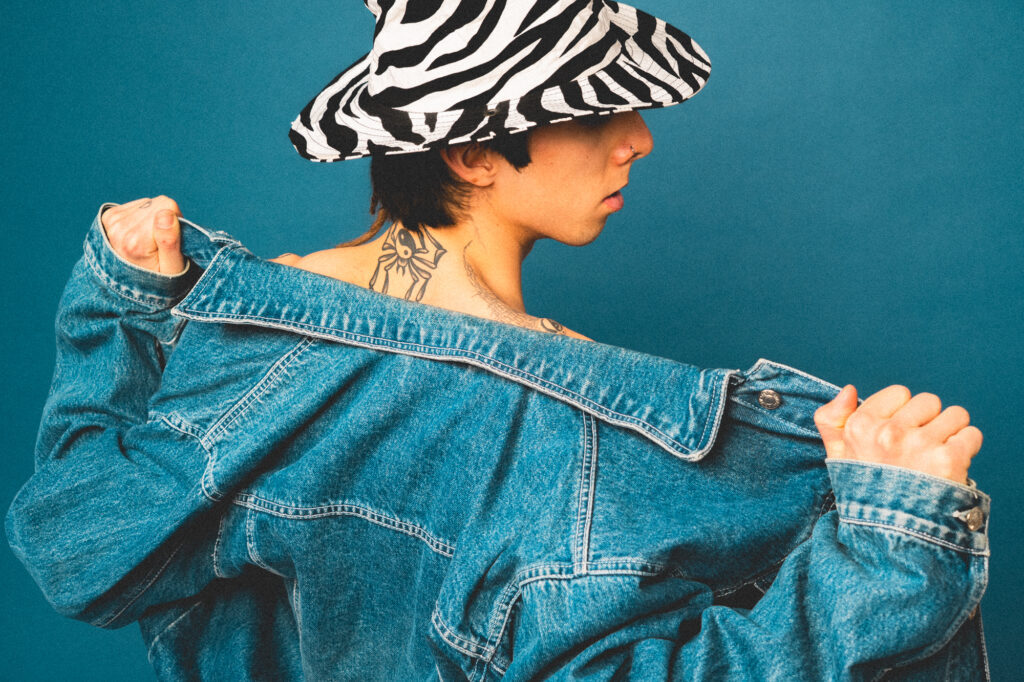 Commercial fashion photography by Łukasz Zając, model in zebra print hat and denim jacket showing neck tattoo against blue background, Poland